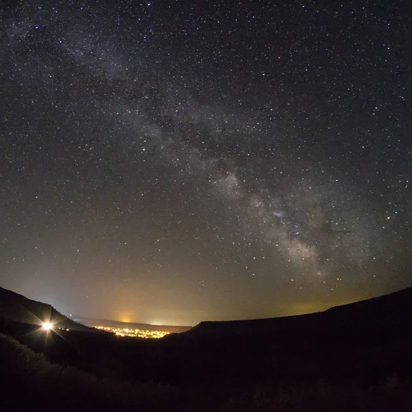 Milky Way above valley with light pollution from a nearby small town and a single light from afar coming up the hill