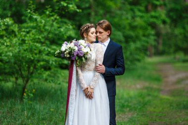 Genç güzel koyu mavi takım elbise ve beyaz kırpma en iyi elbise ve büyük düğün buket Lila ve uzun şeritler tutan gelin damat. Bahar Düğün yürüyüşte mutlu çift