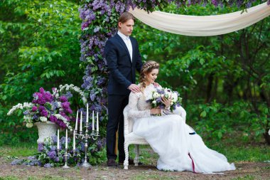 Genç güzel koyu mavi takım elbise ve beyaz kırpma en iyi elbise ve büyük düğün buket Lila ve uzun şeritler tutan gelin damat. Bahar Düğün yürüyüşte mutlu çift