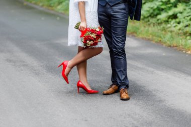 Wedding details: stylish red and brown shoes of bride and groom. Newlyweds standing in front of each other. Bridal bouquet of roses in the hands clipart