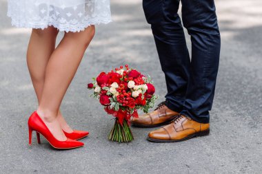 Wedding details: stylish red and brown shoes of bride and groom. Bouquet of roses standing on the ground between them. Newlyweds standing in front of each other clipart