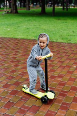 Yağmurda sırılsıklam, bir çocuk bir sporda paten bir scooter üzerinde takım elbise. Bahar yürüyüşte şehir Park, yağmurlu hava.