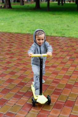 Yağmurda sırılsıklam, bir çocuk bir sporda paten bir scooter üzerinde takım elbise. Bahar yürüyüşte şehir Park, yağmurlu hava.