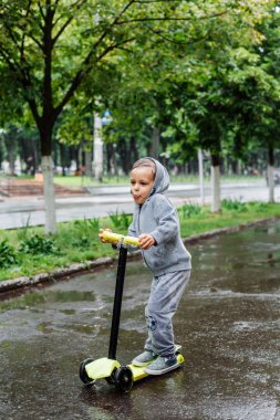 Yağmurda sırılsıklam, bir çocuk bir sporda paten bir scooter üzerinde takım elbise. Bahar yürüyüşte şehir Park, yağmurlu hava.