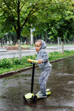 Yağmurda sırılsıklam, bir çocuk bir sporda paten bir scooter üzerinde takım elbise. Bahar yürüyüşte şehir Park, yağmurlu hava.