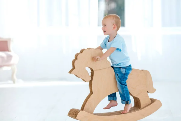 Mignon, souriant, blanc, garçon de deux ans en t-shirt bleu et jean basculant sur un cheval en bois fait main. Petit enfant s'amusant avec un jouet de poney. Concept d'éducation de la petite enfance, famille heureuse — Photo