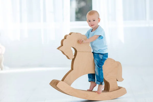 Cute, smiling, white, two years old boy in blue t-shirt and jeans rocking on wooden handmade horse. Little child having fun with pony toy. Concept of early childhood education, happy family
