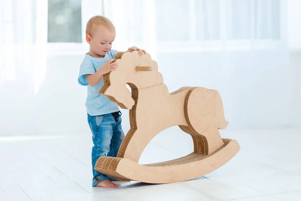 Cute, smiling, white, two years old boy in blue t-shirt and jeans rocking on wooden handmade horse. Little child having fun with pony toy. Concept of early childhood education, happy family