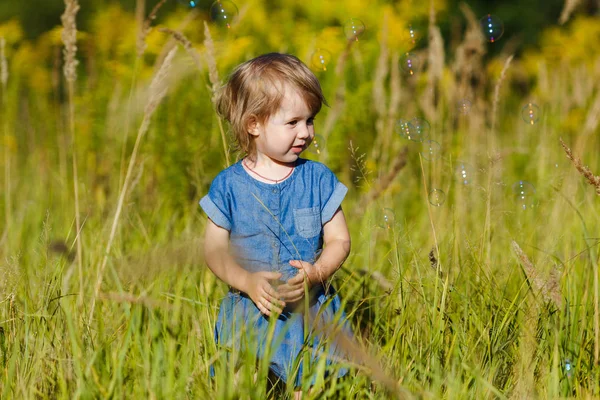 Una ragazzina in abito blu che cerca di prendere bolle di sapone nel parco estivo. Divertente un bambino di un anno che gioca all'aperto. Concetto di vacanze in famiglia e passeggiate nella natura, genitorialità e infanzia felice . — Foto Stock