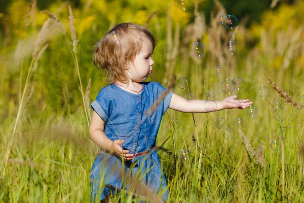 Una ragazzina in abito blu che cerca di prendere bolle di sapone nel parco estivo. Divertente un bambino di un anno che gioca all'aperto. Concetto di vacanze in famiglia e passeggiate nella natura, genitorialità e infanzia felice . — Foto Stock