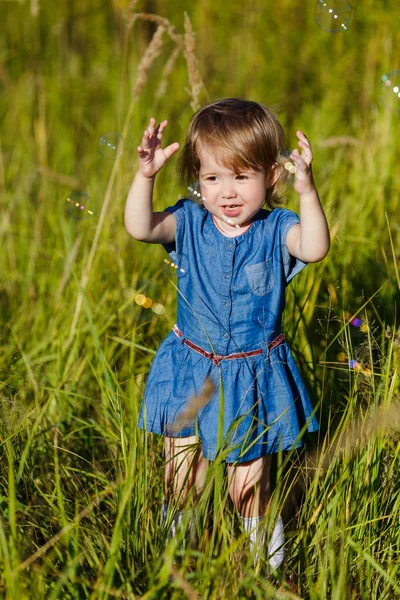 Una ragazzina in abito blu che cerca di prendere bolle di sapone nel parco estivo. Divertente un bambino di un anno che gioca all'aperto. Concetto di vacanze in famiglia e passeggiate nella natura, genitorialità e infanzia felice . — Foto Stock