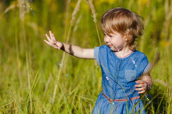 Una ragazzina in abito blu che cerca di prendere bolle di sapone nel parco estivo. Divertente un bambino di un anno che gioca all'aperto. Concetto di vacanze in famiglia e passeggiate nella natura, genitorialità e infanzia felice . — Foto Stock