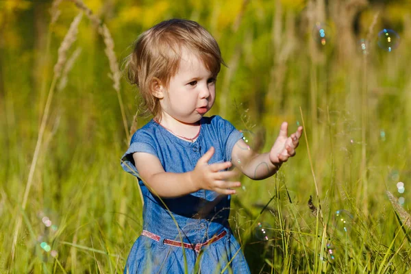 Una ragazzina in abito blu che cerca di prendere bolle di sapone nel parco estivo. Divertente un bambino di un anno che gioca all'aperto. Concetto di vacanze in famiglia e passeggiate nella natura, genitorialità e infanzia felice . — Foto Stock