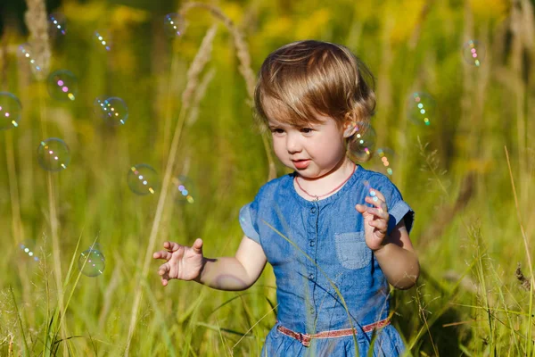 Una ragazzina in abito blu che cerca di prendere bolle di sapone nel parco estivo. Divertente un bambino di un anno che gioca all'aperto. Concetto di vacanze in famiglia e passeggiate nella natura, genitorialità e infanzia felice . — Foto Stock