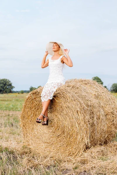 Pięknie Opalona dziewczyna w białej sukni koronki jest stojący w pobliżu stogu siana o zachodzie słońca w lecie. Szczęśliwa, uśmiechnięta kobieta w słomkowy kapelusz, dobra zabawa, pozowanie na polu. Koncepcja życia wsi. — Zdjęcie stockowe
