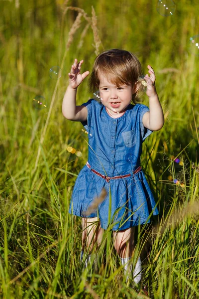 A little girl in blue dress trying to catch soap bubbles in summer park. Funny one year old kid playing outdoors. Concept on family holidays and walking at nature, parenting and happy childhood. Royalty Free Stock Photos