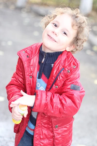 Lockigt gladlynt pojke i en ljust röd jacka som går på gatan i höst och blåsa bubblor — Stockfoto