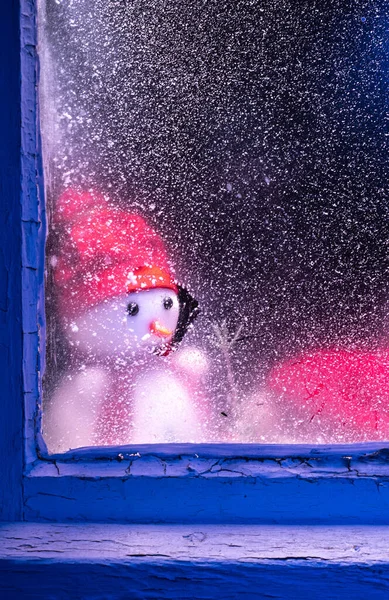 Ένας θλιμμένος χιονάνθρωπος κοιτάζει από το παράθυρο. Παράθυρο ακόμα φόντο με πάγο και χιόνι για τη διακόσμηση και το χειμερινό τοπίο του δάσους με χιονάνθρωπο. Χριστούγεννα και ηλιόλουστη κρύα μέρα. — Φωτογραφία Αρχείου
