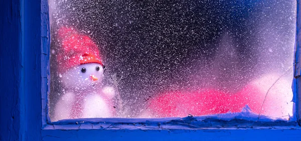 Smutný sněhulák se dívá oknem. Okno ještě pozadí s ledem a sněhem pro dekoraci a zimní krajinu lesa se sněhulákem. Vánoční čas a slunečný chladný den. Stock Snímky
