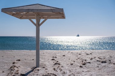 Kumsal. Güneşten gelen şemsiye. Kum. Tepe örtüsü. Deniz. Mavi gökyüzü. Kimse yok.. 