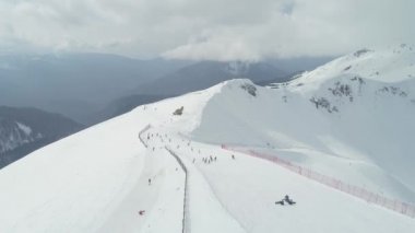 Kayak merkezi. Dağ yamacında kayakçılar ve snowboardcular. Yüksek karlı dağlar. Krasnaya Polyana, Soçi. Hava fotoğrafçılığı videosu. Kar yağıyor. Bulutlu. Sezon.