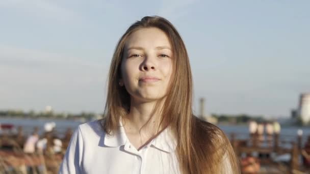 Mouvement lent du portrait de la jeune femme blonde regardant dans la caméra, sourire mignon, soleil brillant, fond de rivière. Mignon modèle de visage souriant. La fille regarde la caméra avec joie. Blond cheveux mouche vent battant — Video