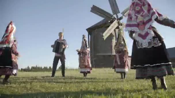 伝統的なロシアの服の人々は、伝統的なアンティークの木製の風車の背景に屋外で踊り、歌を歌います。国民服を着た幸せな人々のグループ｜Finno-Ugric服. — ストック動画