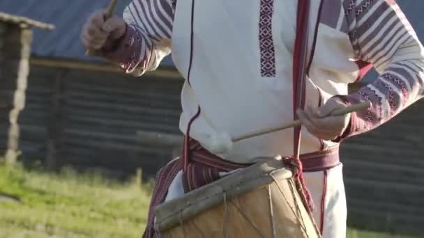 伝統的なロシアの服の男は、伝統的なアンティークの木製の風車の背景に屋外で踊り、民俗ドラムで演奏します。民族衣装を着た男｜Finno-Ugric服. — ストック動画
