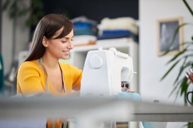 Gülümseyen beyaz tenli moda tasarımcısı stüdyosunda oturuyor ve güzel bir gece elbisesi dikiyor..