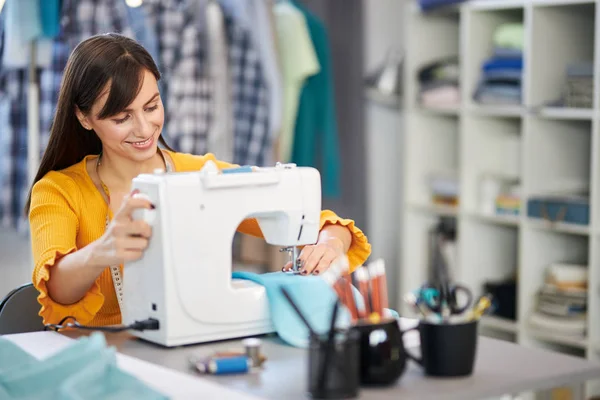 Sonriendo encantadora diseñadora de moda caucásica sentada en su estudio y cosiendo hermoso vestido de noche . —  Fotos de Stock