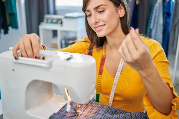 Souriante couturière dévouée assise dans son studio et insérant du fil sur une machine à coudre . — Photo