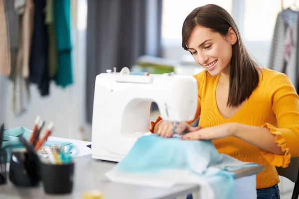 Smiling charming caucasian fashion designer sitting in her studio and sewing beautiful evening dress. — Stock Photo, Image