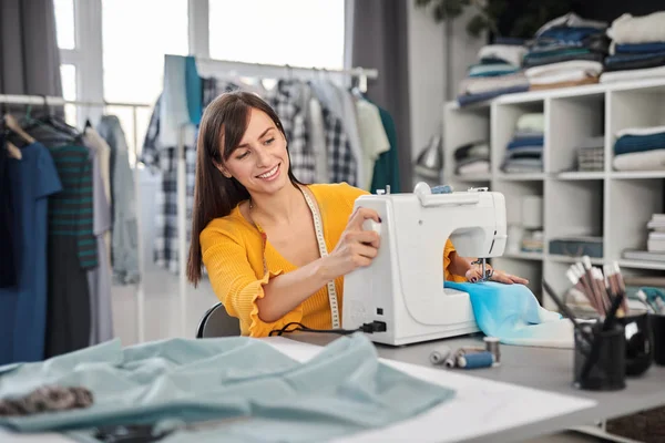 Smiling charming caucasian fashion designer sitting in her studio and sewing beautiful evening dress. — Stock Photo, Image
