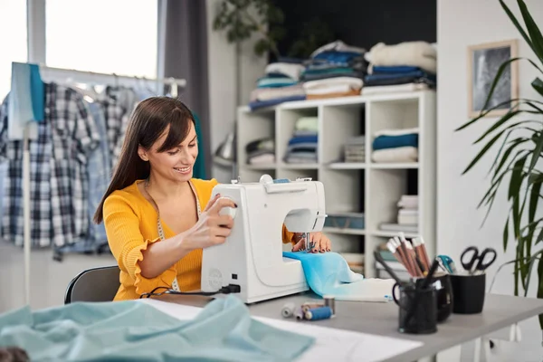 Sonriendo encantadora diseñadora de moda caucásica sentada en su estudio y cosiendo hermoso vestido de noche . —  Fotos de Stock