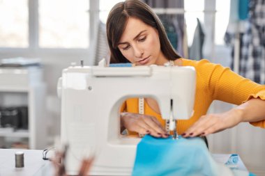 Focused charming caucasian fashion designer sitting in her studio and sewing beautiful evening dress. clipart