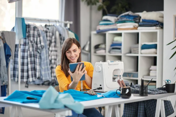 Sorrindo positivo caucasiano morena sentada em seu estúdio de moda e usando tablet . — Fotografia de Stock