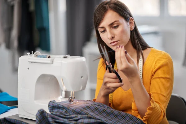 Attractive smiling caucasian brunette sitting in tailoring studio and cutting thread. — Stockfoto