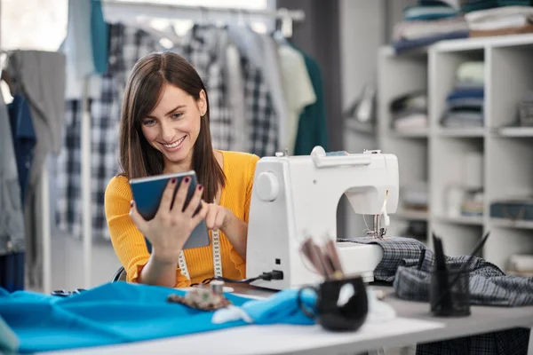 Sorrindo positivo caucasiano morena sentada em seu estúdio de moda e usando tablet . — Fotografia de Stock