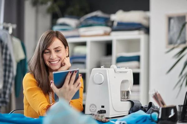 Sonriente morena caucásica positiva sentada en su estudio de moda y usando la tableta . —  Fotos de Stock