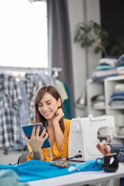 Gülümseyen, beyaz tenli esmer, moda stüdyosunda oturuyor ve tablet kullanıyor..