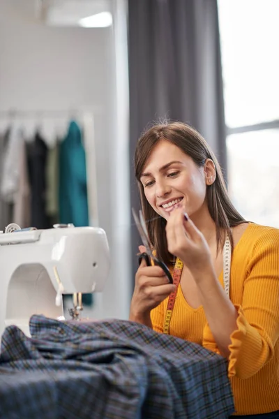 Attractive smiling caucasian brunette sitting in tailoring studio and cutting thread. — Stock Photo, Image