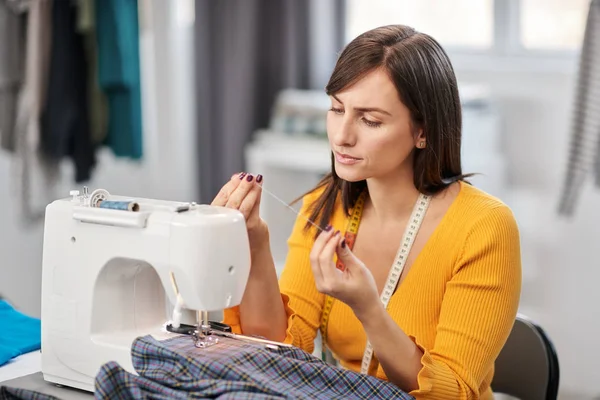 Costurera dedicada sentada en su estudio e insertando hilo en la máquina de coser . —  Fotos de Stock