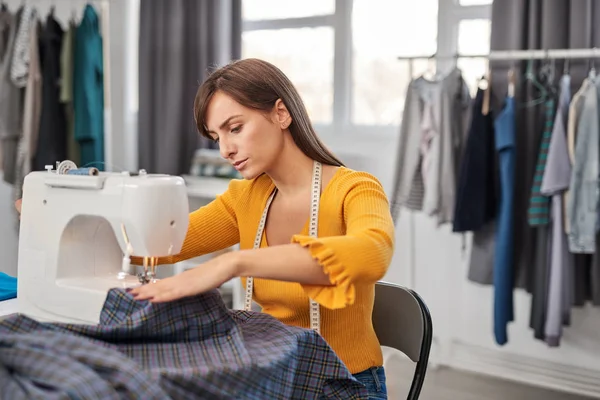Focused charming caucasian fashion designer sitting in her studio and sewing beautiful dress. — Stock Photo, Image