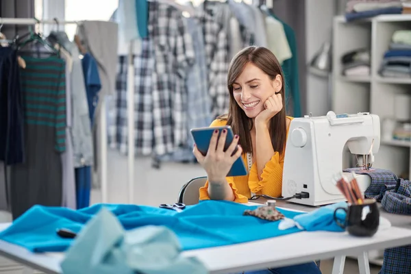 Sorridente positivo caucasico bruna seduta nel suo studio di moda e utilizzando tablet . — Foto Stock