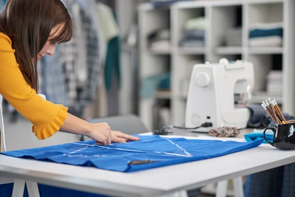 Creative caucasian fashion designer standing in her studio and drawing scheme on blue linen for a beautiful evening dress. — Stock Photo, Image