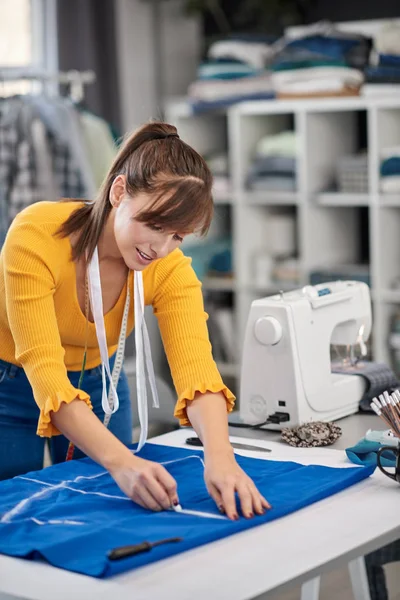 Creative caucasian fashion designer standing in her studio and drawing scheme on blue linen for a beautiful night dress. — Stock Photo, Image