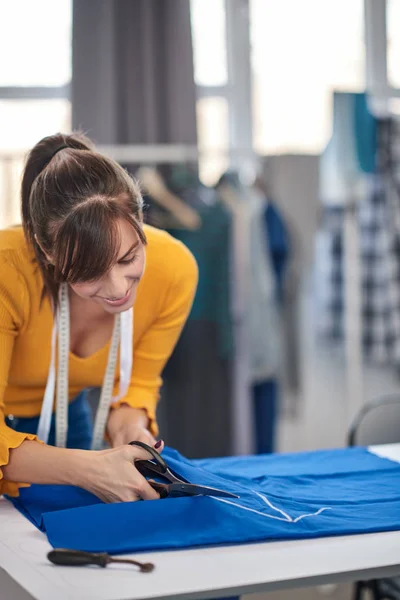 Costurera dedicada de pie junto a la mesa y corte de textiles para un vestido elegante . —  Fotos de Stock