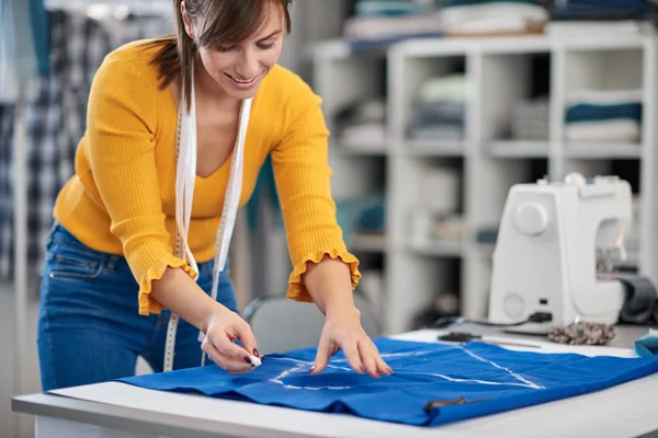 Creative caucasian fashion designer standing in her studio and drawing scheme on blue linen for a beautiful night dress. — Stock Photo, Image