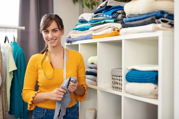 Belle souriante couturière caucasienne tenant morceau de textile et debout à côté de l'étagère avec des vêtements dans son studio . — Photo