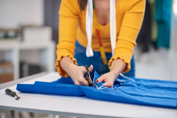 Costurera dedicada de pie junto a la mesa y corte de textiles para un vestido elegante . — Foto de Stock